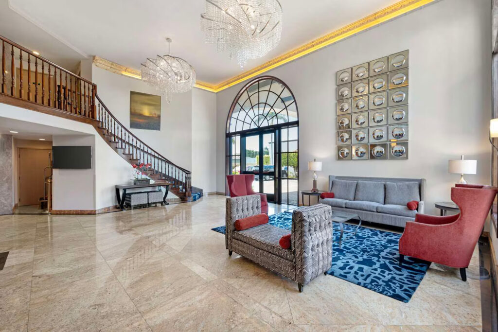 Modern hotel lobby at Best Western Costa Mesa with marble floors, a curved wooden staircase, large arched windows, gray sofas, red chairs, blue patterned rug, two chandeliers, and contemporary wall art enhancing the inviting space.