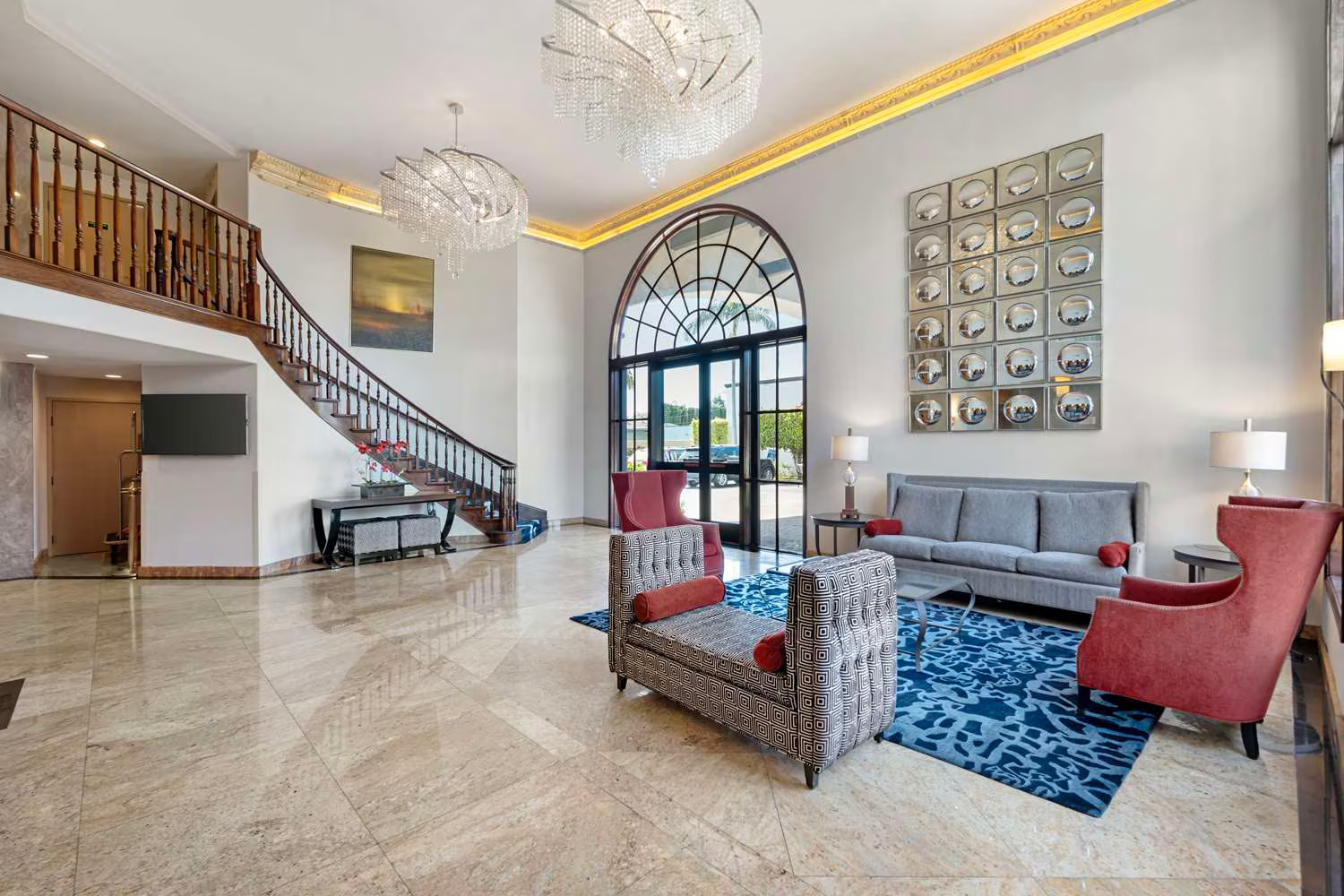 Modern hotel lobby at Best Western Costa Mesa with marble floors, a curved wooden staircase, large arched windows, gray sofas, red chairs, blue patterned rug, two chandeliers, and contemporary wall art enhancing the inviting space.
