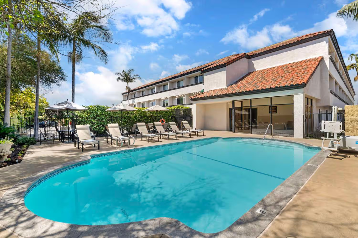 Enjoy the outdoor hotel pool at Best Western Costa Mesa, with clear blue water, lounge chairs, and umbrellas. Palm trees and greenery surround a three-story building with red roof tiles under a partly cloudy sky.
