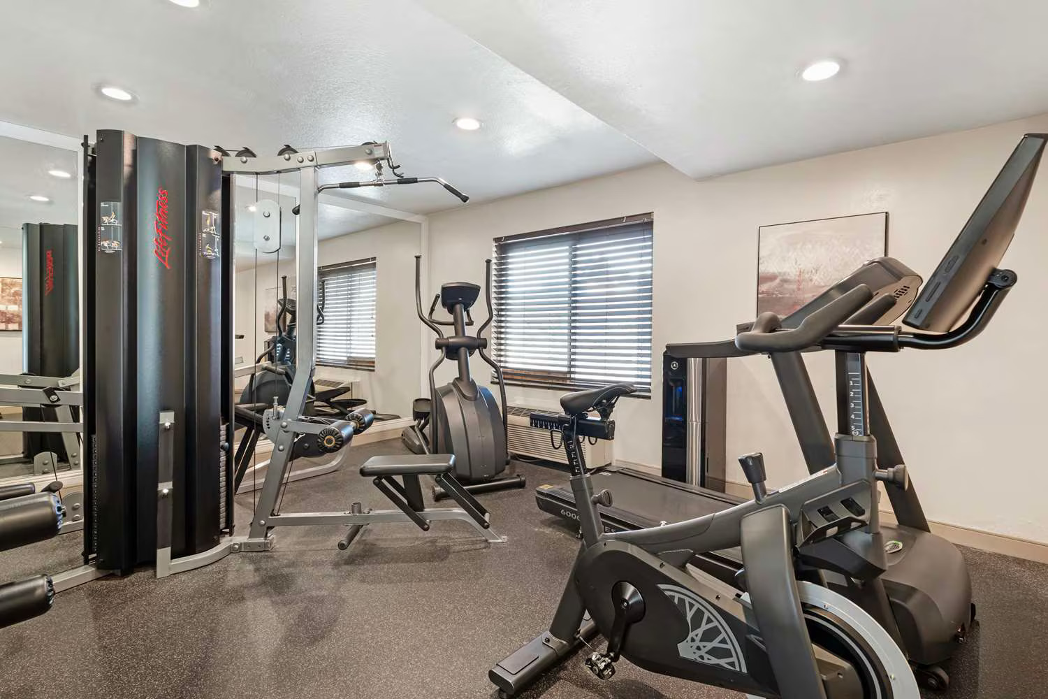 A modern home gym at Best Western Costa Mesa features a treadmill, elliptical machine, weight bench, and multi-function weight station. Large windows with blinds, neutral walls, bright lighting, and a framed picture create an inviting space.