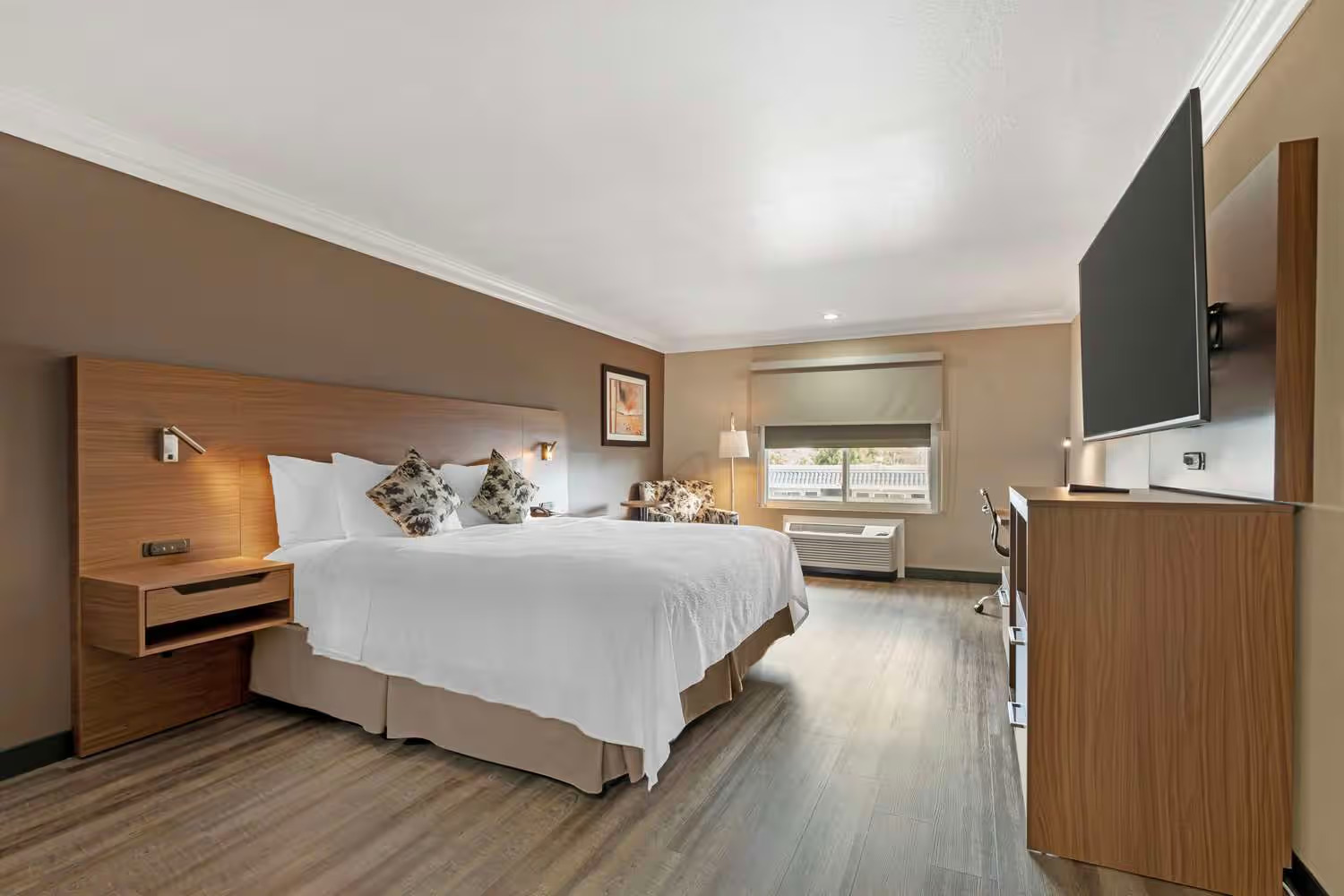 A spacious Best Western Costa Mesa hotel room featuring a large bed, two bedside tables, a wall-mounted TV, a desk with chair, window with blinds, and wooden floors. The modern decor showcases neutral tones for a relaxing stay.
