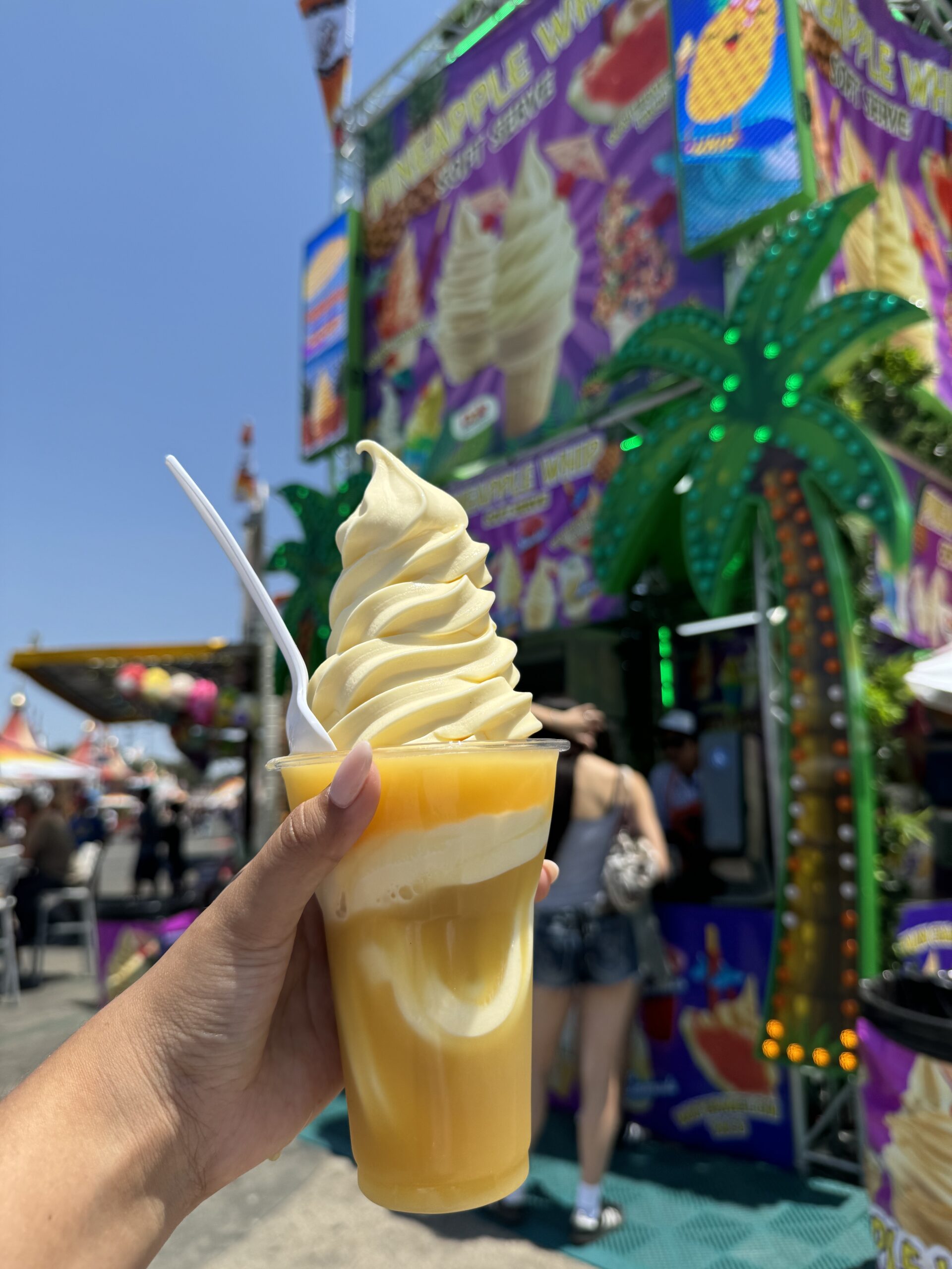 A hand holds a cup with swirled soft-serve ice cream and yellow slush, complete with a white spoon and straw. In the background, the lively OC Fair Costa Mesa food stand features tropical signs and a glowing palm tree.