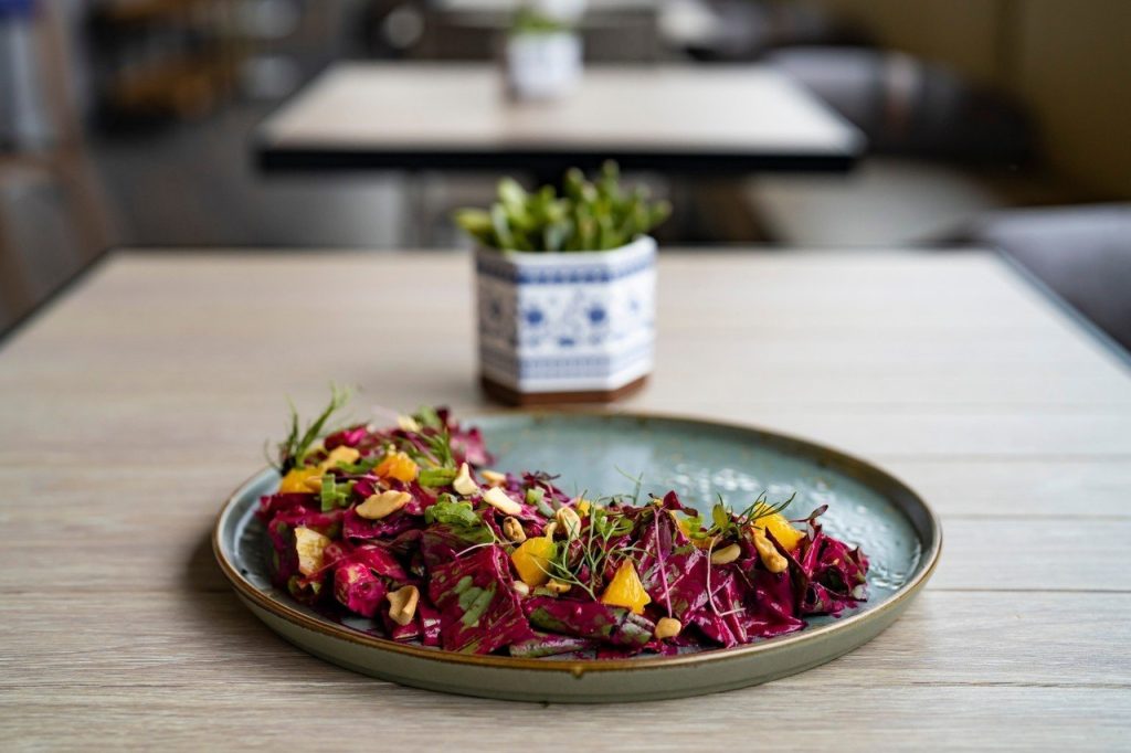 A plate of vibrant salad with red cabbage, yellow vegetables, greens, and sliced almonds sits on a wooden table; a small potted plant is blurred in the background.