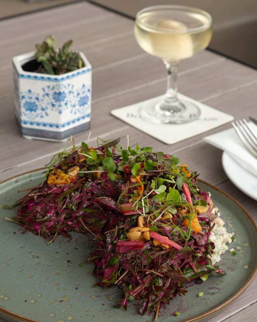 A plate of colorful salad with microgreens and vegetables sits on a wooden table next to a glass of white wine, a blue and white potted plant, and a fork and knife on a napkin.