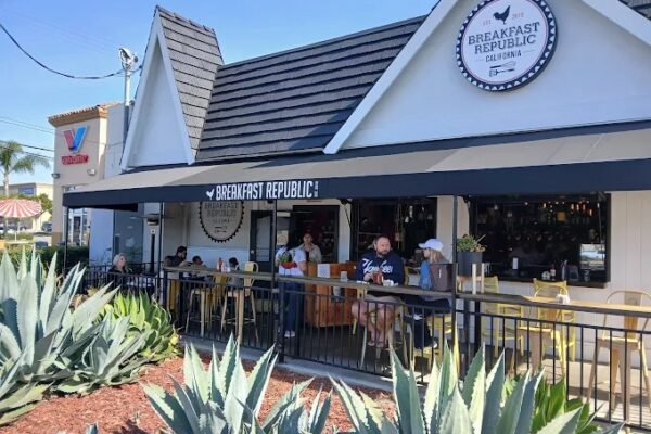 People dine on the outdoor patio of Breakfast Republic Costa Mesa, a restaurant with a black awning and white exterior. Succulent plants fill the foreground, while a blue sky stretches across the background.