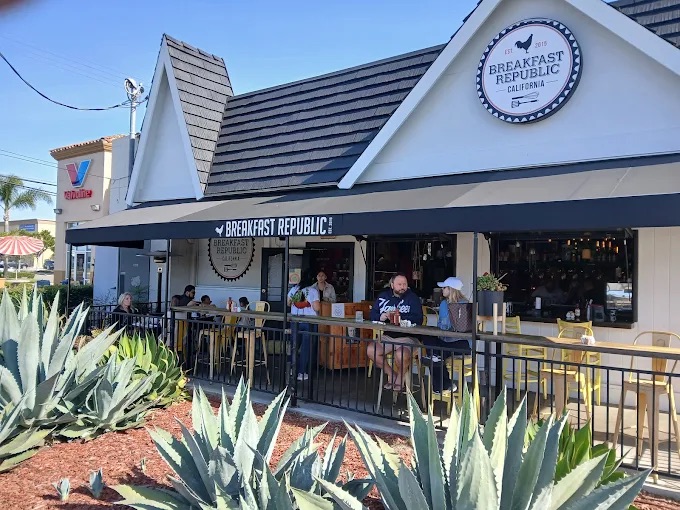 People dine on the outdoor patio of Breakfast Republic Costa Mesa, a restaurant with a black awning and white exterior. Succulent plants fill the foreground, while a blue sky stretches across the background.
