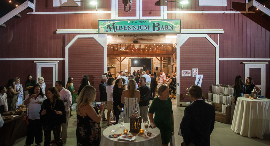 A group of people socialize and mingle at an evening event outside a red barn labeled Millennium Barn, one of the charming meeting venues Costa Mesa offers. Tables with drinks and decorations are set up, while string lights hang inside the barn entrance.