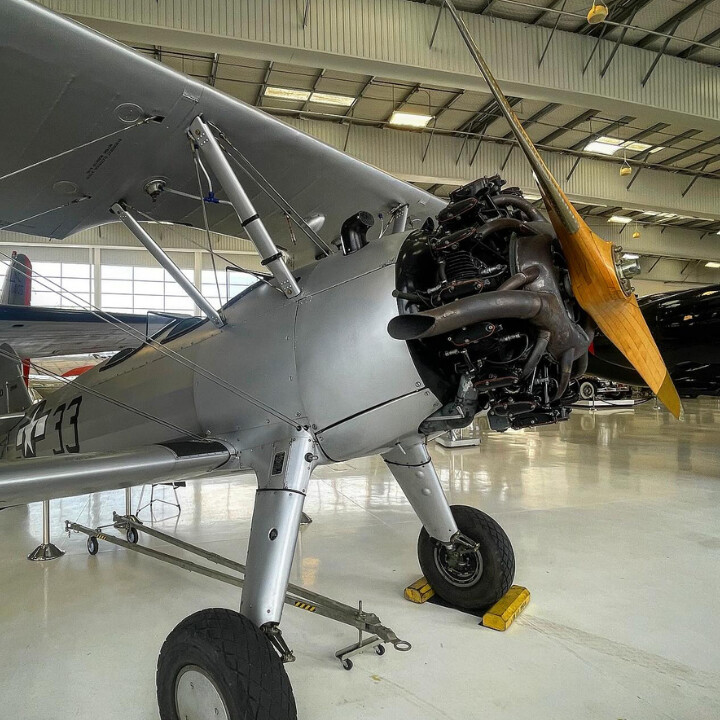 A vintage silver biplane with a wooden propeller is displayed in a brightly lit Costa Mesa hangar. The aircraft’s exposed engine and landing gear are visible, with other planes showcased in the background.