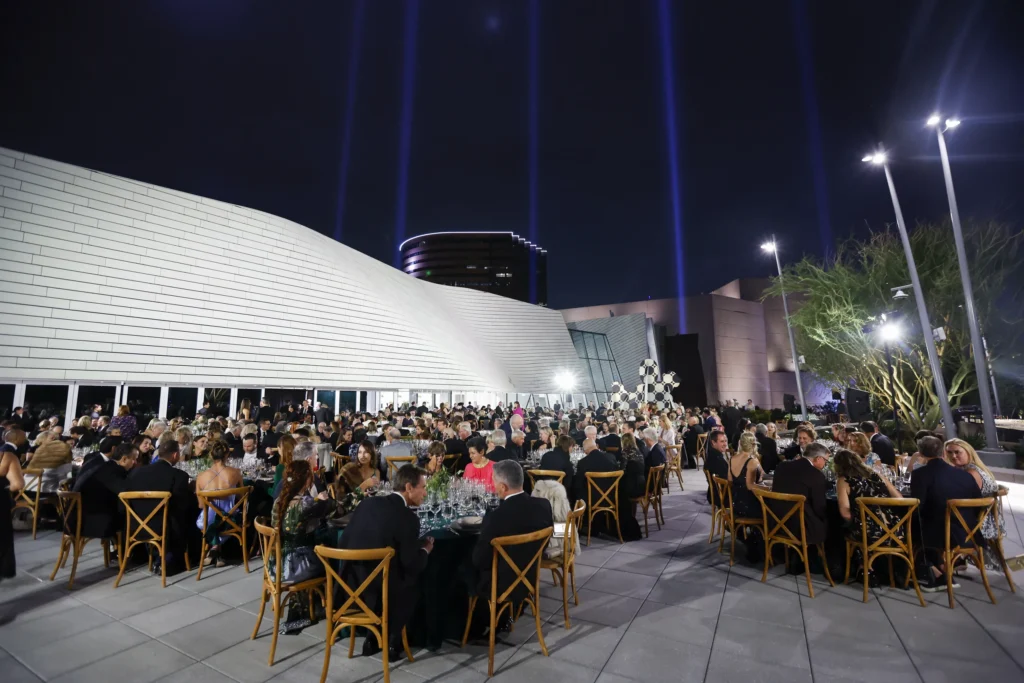 A large group of people dressed formally are seated at round tables outdoors at night, attending an event with a modern white building and dramatic light beams—showcasing why event venues in Costa Mesa are so sought after.
