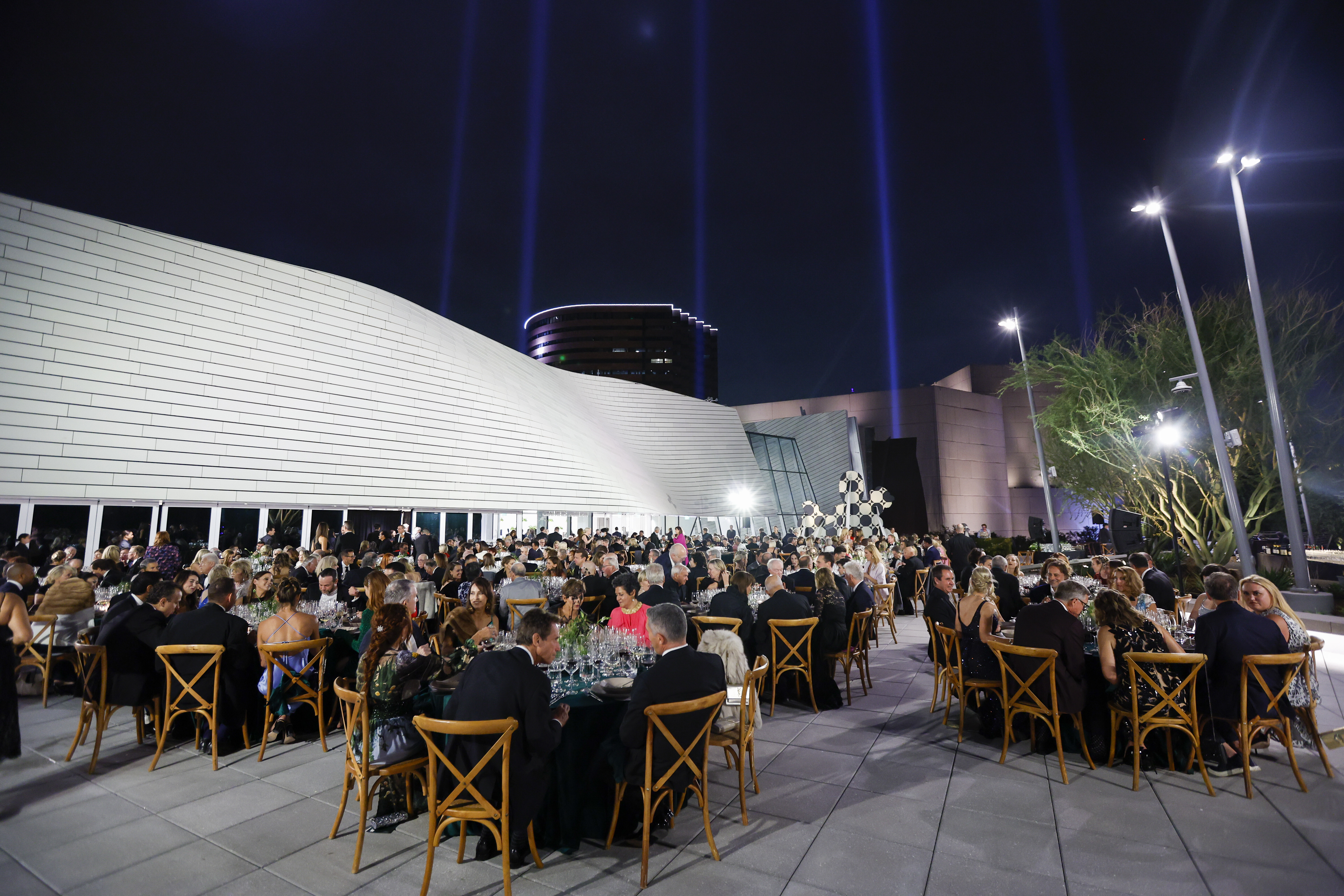 A large group of people dressed formally are seated at round tables outdoors at night, attending an event with a modern white building and dramatic light beams—showcasing why event venues in Costa Mesa are so sought after.