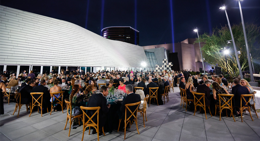 A large group of people dressed formally are seated at round tables outdoors at night, attending an elegant event beside a modern white building—one of the premier meeting venues Costa Mesa has to offer, featuring striking architectural lighting.