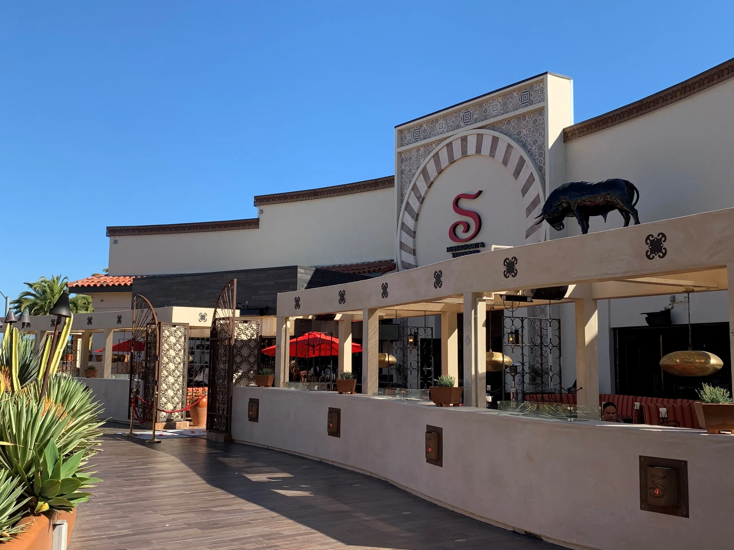 The image shows the exterior of a restaurant at Triangle Square Costa Mesa, with Mediterranean-style architecture, a large S logo above the entrance, decorative tiles, outdoor seating, red umbrellas, and a bull sculpture on the roof.