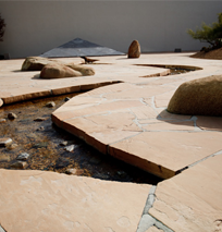 Tan stone slabs form a geometric pathway over a shallow stream, with large rocks and sparse greenery in a minimalist space—evoking the modern flair of a costa mesa art walk, bordered by a plain wall.