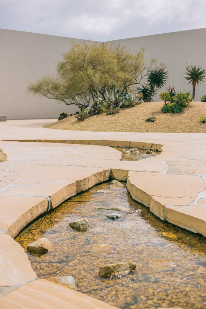 A small stream with clear water flows through a desert garden, bordered by light stone slabs, reminiscent of the Noguchi Garden Costa Mesa. In the background are cactus plants, dry grass, and a leafless tree against a high white wall.