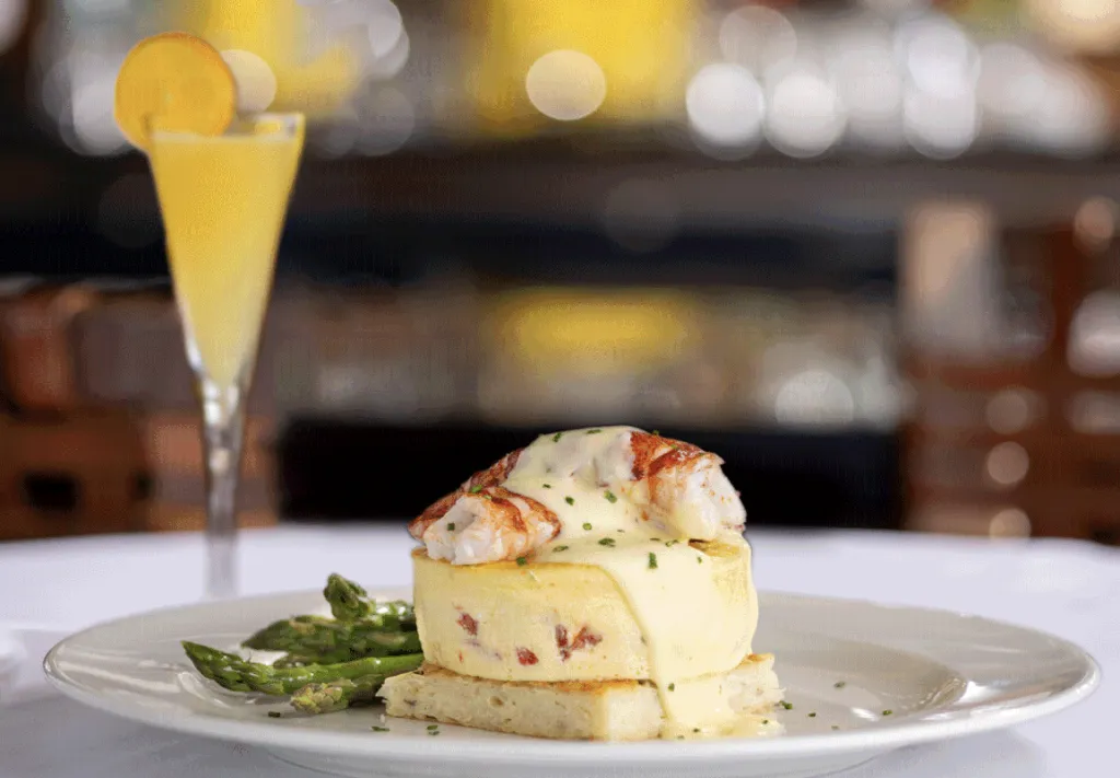A plate with a savory egg dish topped with creamy sauce and lobster, served with asparagus. In the background, there is a glass of a yellow beverage with a lemon slice on the rim.