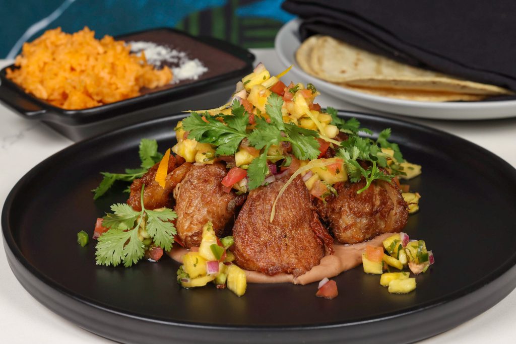 Crispy pieces of meat topped with fresh salsa and cilantro served on a black plate, with a side of shredded orange vegetable and tortillas in the background.