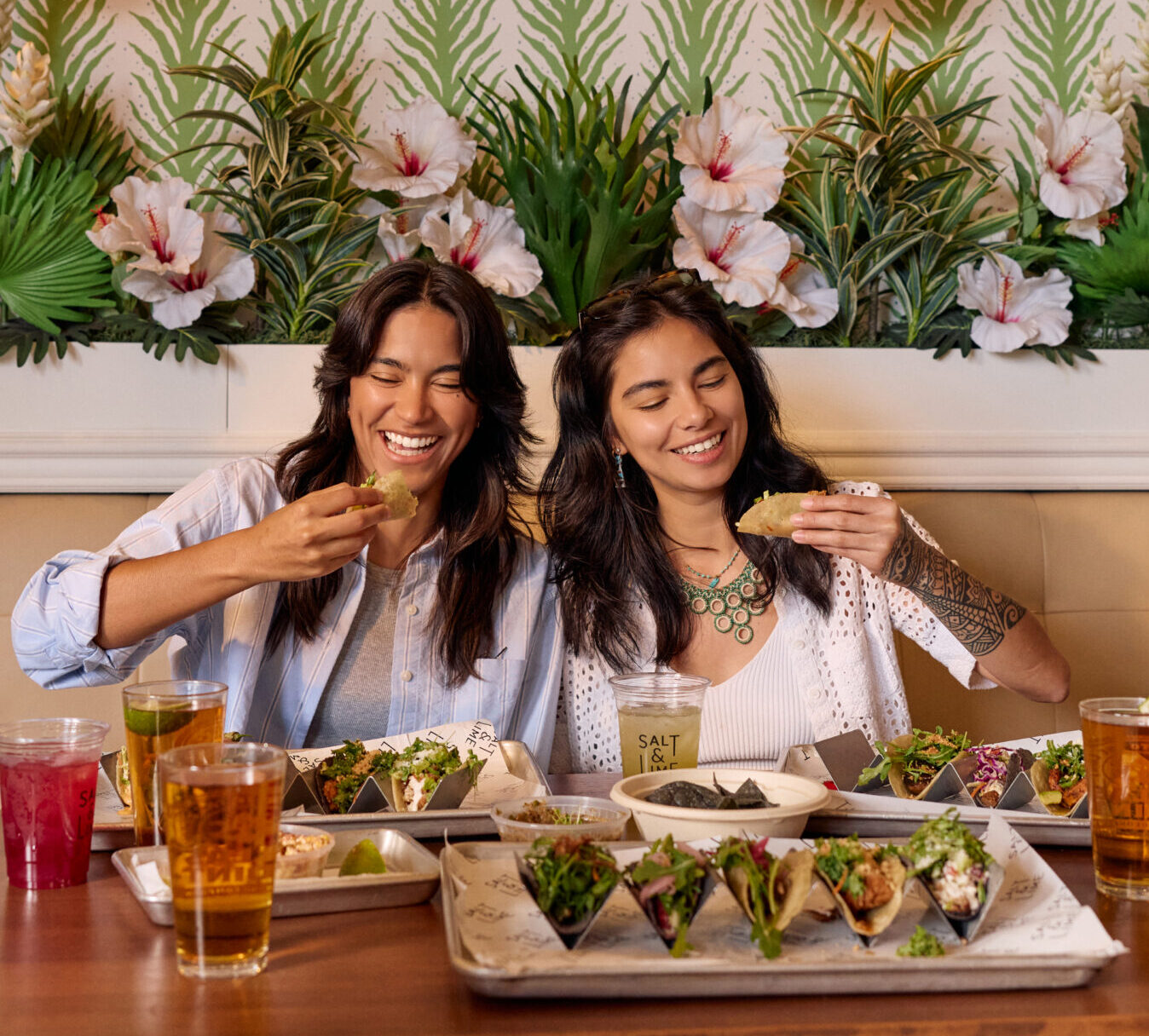 Two women sit at a table in Costa Mesa, enjoying tacos and drinks, smiling and laughing. Various food trays and beverages are spread before them. The background features a decorative wall with green leaves and large white hibiscus flowers.
