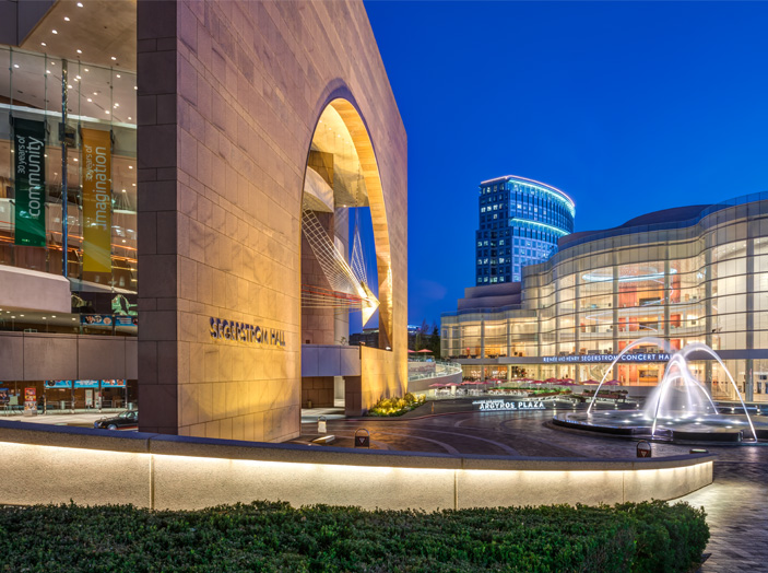 A brightly lit performing arts center in Costa Mesa at dusk features Segerstrom Hall’s grand archway, a modern glass building, tall banners, and a fountain with water jets in the plaza. A cityscape with an office tower is visible in the background.