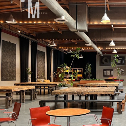 A spacious, modern room with wooden tables, red chairs, potted plants, industrial lighting, and a high ceiling with exposed ductwork—perfect for meeting venues Costa Mesa. Large artworks adorn the walls and bookshelves line the background.