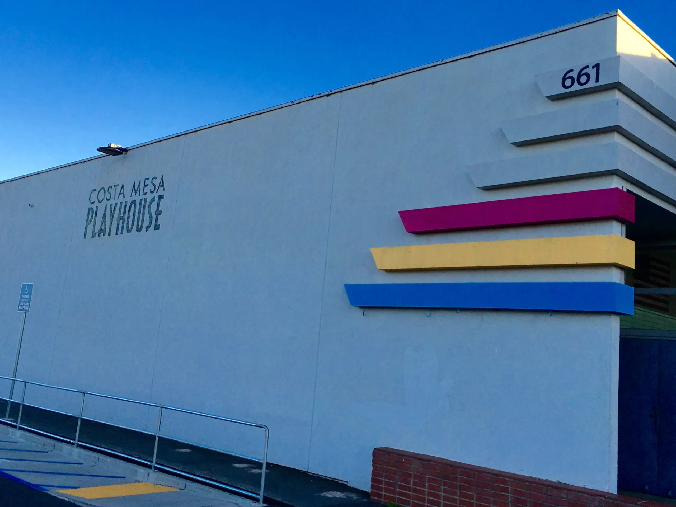 Exterior of the Costa Mesa Playhouse at 661, featuring a white wall with pink, yellow, and blue horizontal accents.