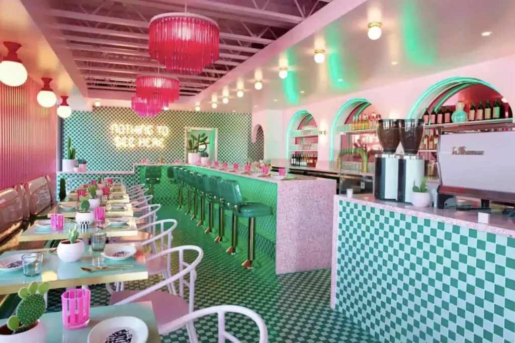 A retro-style diner with green and white checkered floors and counters, pink lighting, bar stools, white chairs, and a neon sign that reads ORDER TO RECEIVE.