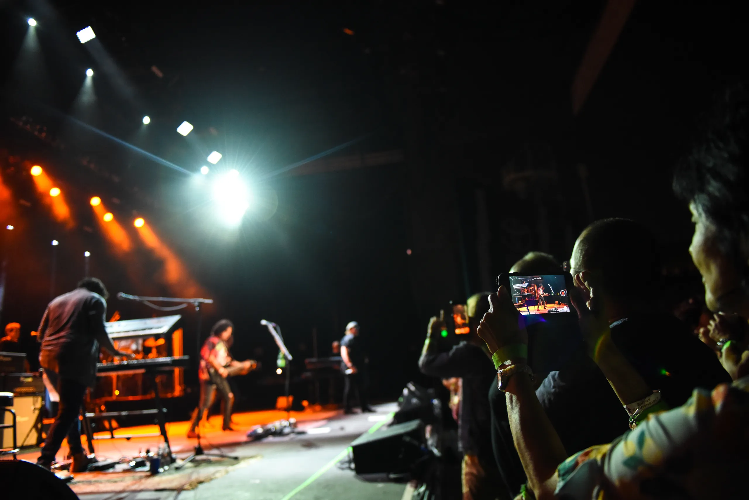 A crowd watches a live concert on a dark stage with orange lights; one person in the audience is recording the performance on a smartphone.