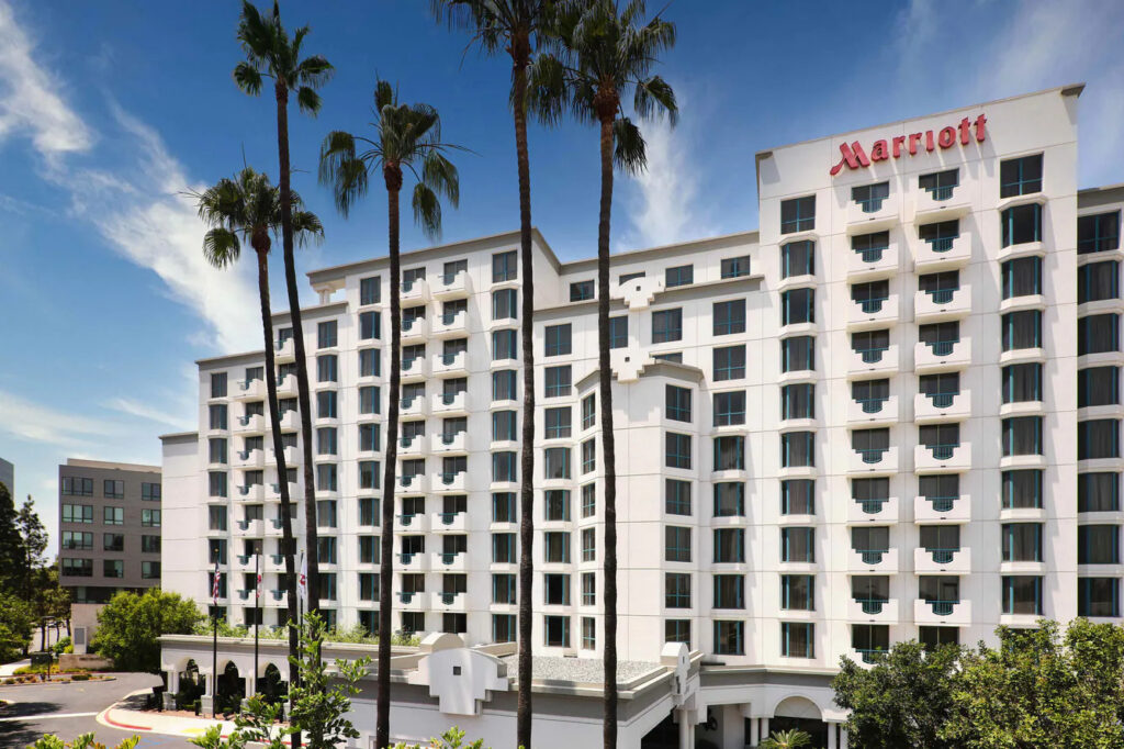 A large white Marriott Costa Mesa hotel building with multiple balconies, tall palm trees in front, and a clear blue sky overhead. The hotel entrance and surrounding greenery are visible at the base.