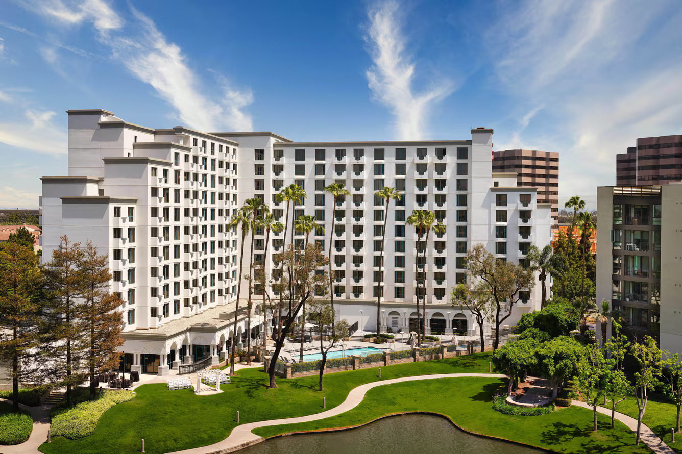 The Marriott Costa Mesa is a large white hotel with balconies overlooking a swimming pool, tall palm trees, and a landscaped garden with a small pond, all under a blue sky with wispy clouds.