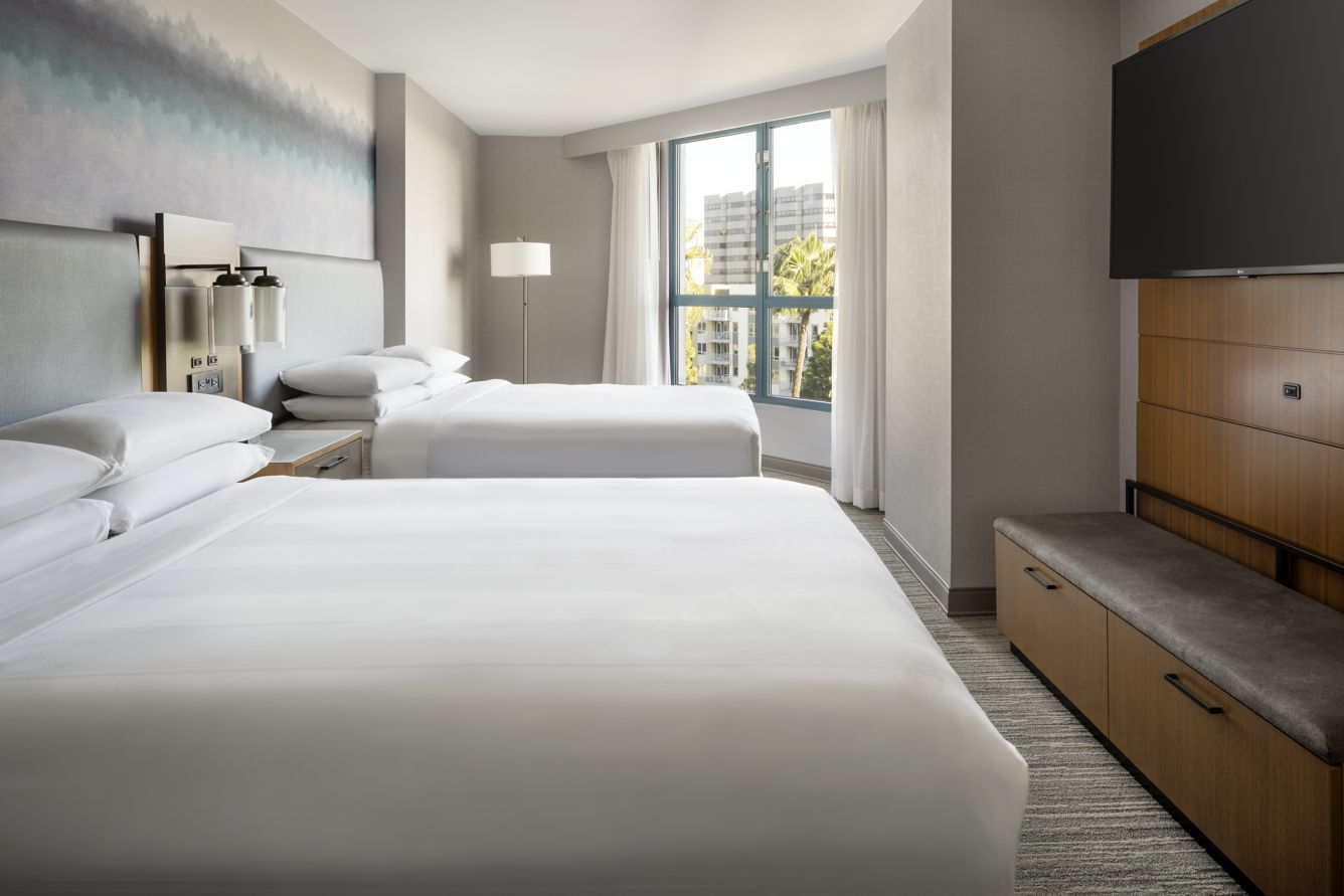 Modern Marriott Costa Mesa hotel room with two large beds, a floor lamp, wall-mounted TV, wooden furniture, and a large window letting in natural light and offering a stunning cityscape view.