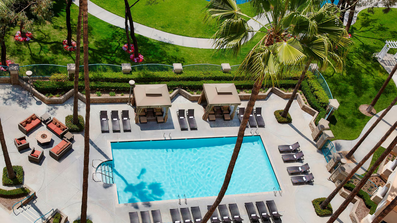 Aerial view of the Marriott Costa Mesa’s rectangular outdoor swimming pool, surrounded by lounge chairs, cabanas, palm trees, and landscaped gardens, with a stylish fire pit seating area nearby.