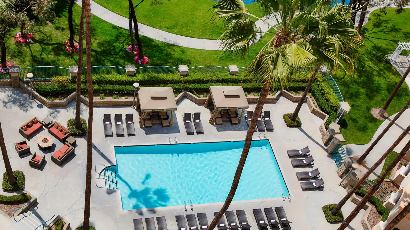 Aerial view of the Marriott Costa Mesa’s rectangular outdoor swimming pool, surrounded by lounge chairs, cabanas, palm trees, and landscaped gardens, with a stylish fire pit seating area nearby.