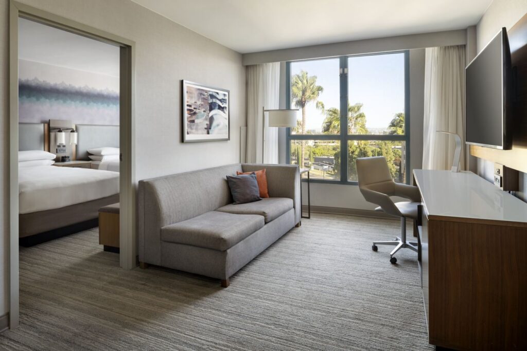 Experience comfort in this modern Marriott Costa Mesa suite, featuring a gray sofa, desk, office chair, large TV, and a window overlooking palm trees. The bedroom with a bed and bedside lamps is visible through an open doorway.