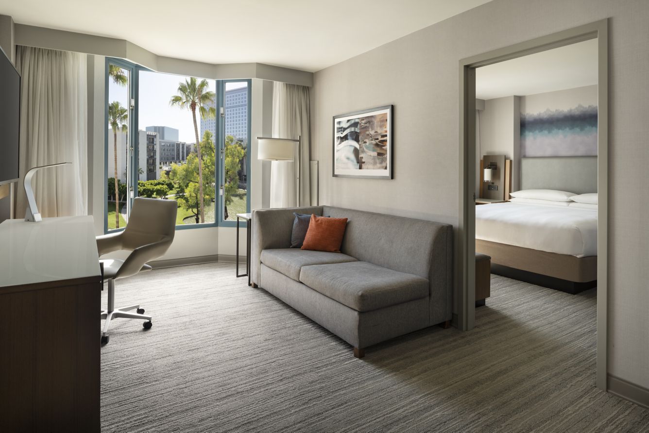Modern Marriott Costa Mesa hotel suite with a gray sofa, desk, and office chair in the living area, large window with palm tree views, and a separate bedroom featuring a king-sized bed and contemporary decor.