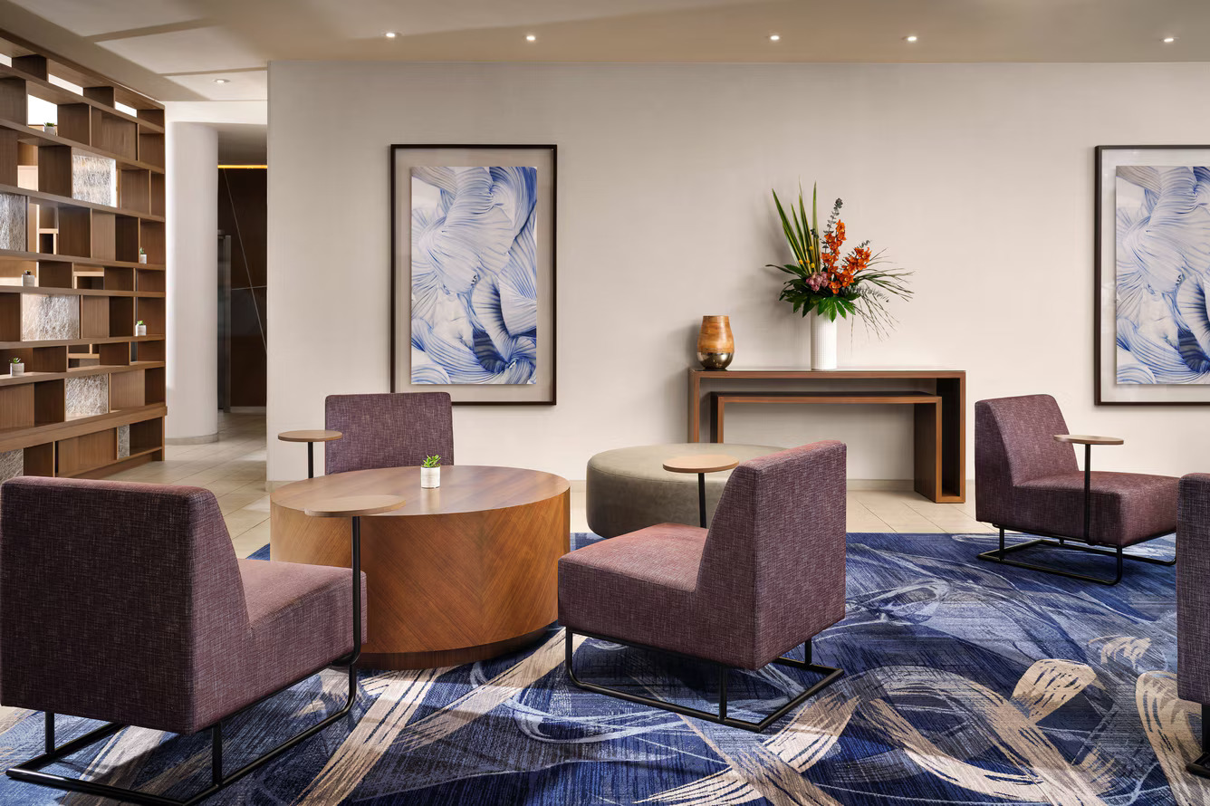 Modern lobby lounge at Marriott Costa Mesa with four purple armchairs around a wooden table, a blue patterned rug, abstract art on the walls, a console table with a floral arrangement, and a decorative wooden shelving unit.
