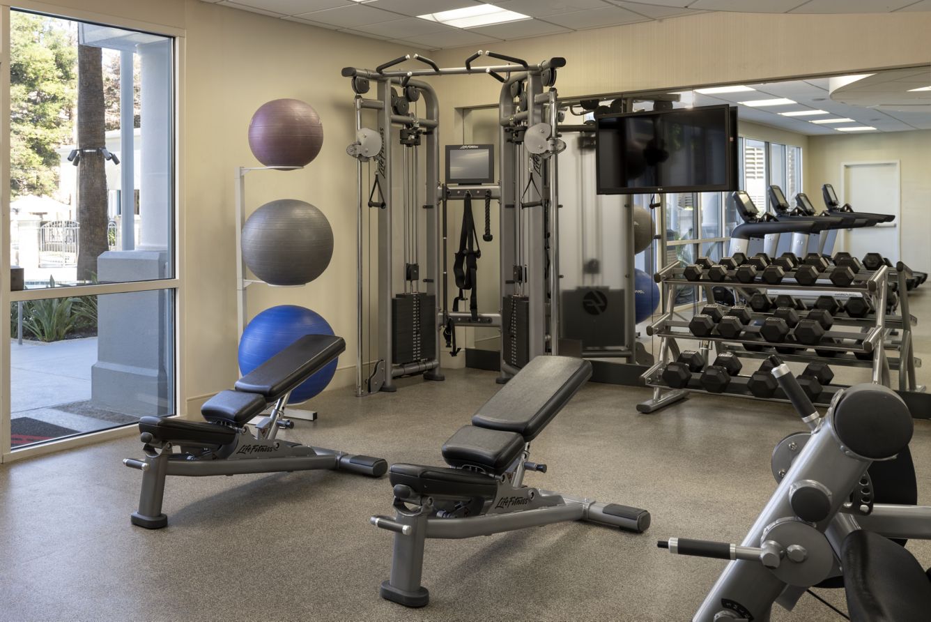 A modern gym at Marriott Costa Mesa features weight benches, dumbbell racks, a cable machine, exercise balls mounted on the wall, and large windows letting in natural light.