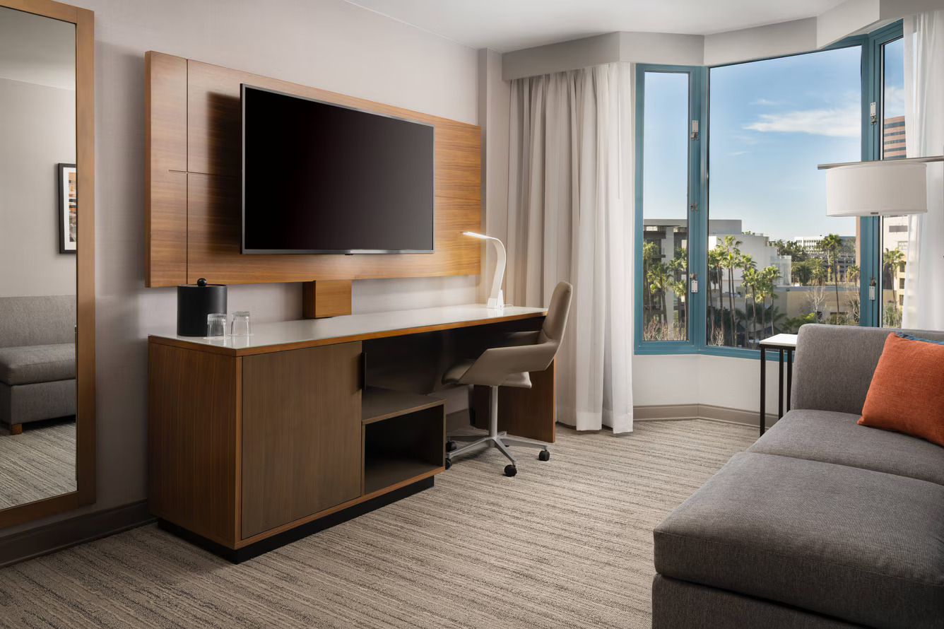 Modern hotel room at Marriott Costa Mesa with a large wall-mounted TV, desk and lamp, cozy sofa with orange cushion, full-length mirror, and a window offering cityscape views with palm trees and blue sky.