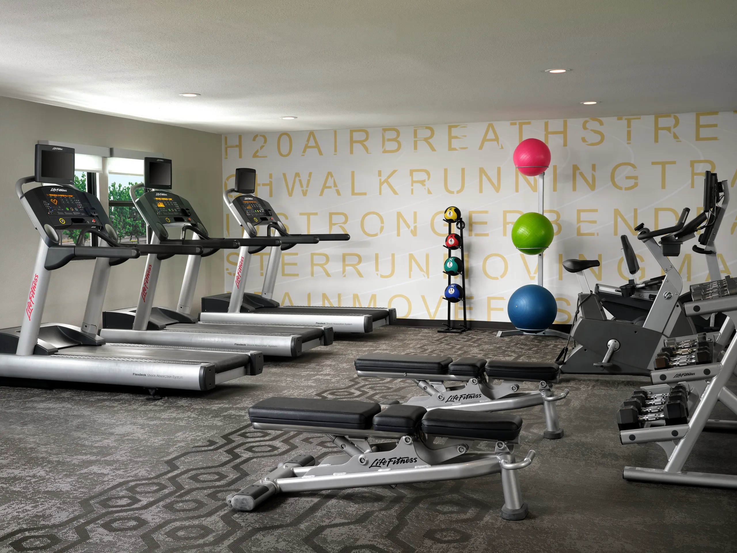 Modern gym at Residence Inn Costa Mesa with treadmills, stationary bikes, benches, colorful exercise balls, and a rack of weights. Yellow words are printed on the back wall. Large windows let in natural light.