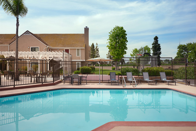A fenced outdoor swimming pool with lounge chairs, surrounded by greenery and trees, sits beside a light-colored house at Residence Inn Costa Mesa under a blue sky with scattered clouds.