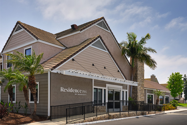 A tan, two-story building with a sloped roof and palm trees sits under a blue sky, its sign reading Residence Inn Costa Mesa by Marriott on the exterior.