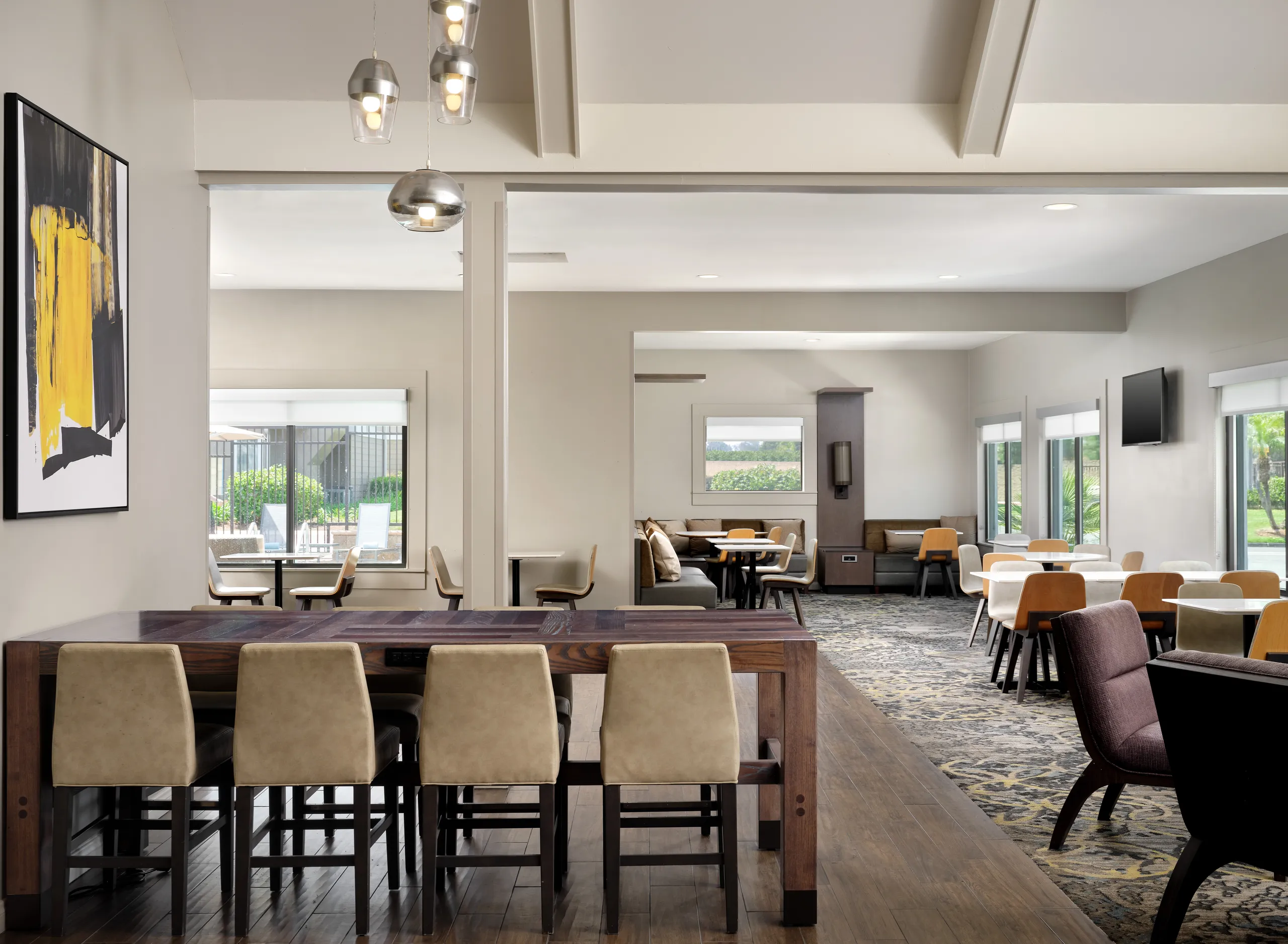Modern dining area at Residence Inn Costa Mesa with beige chairs, wooden tables, large windows, patterned carpet, wall art, and pendant lights in a bright, spacious room.