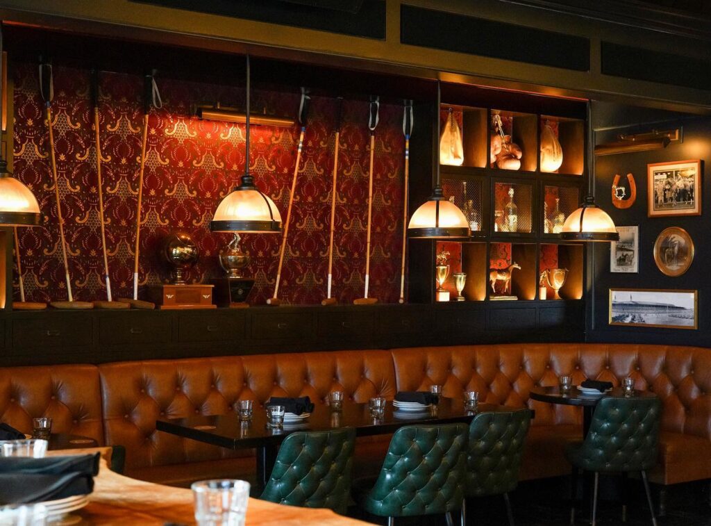 A stylish restaurant interior, perfect for a Costa Mesa travel blog, with orange tufted leather seating, green quilted chairs, dark tables set with plates and glasses, warm lighting, and red patterned wallpaper displaying trophies and framed photos.