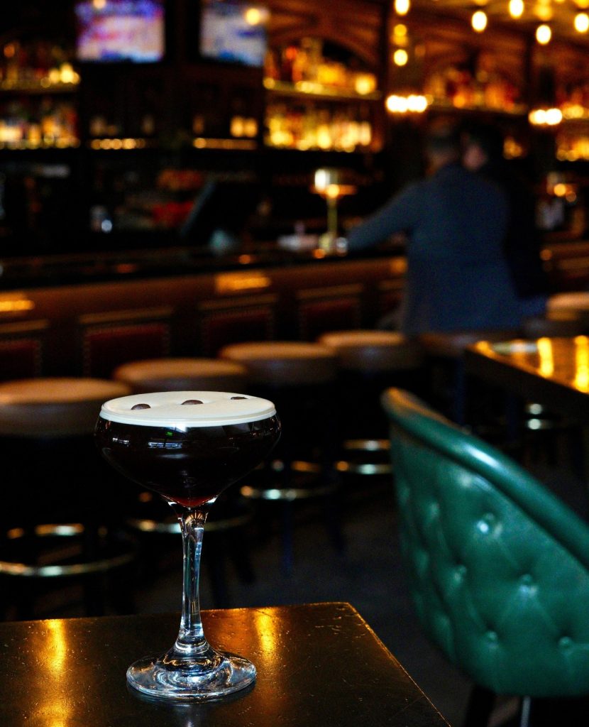 A dark espresso martini topped with three coffee beans sits on a table in a dimly lit bar, with bar stools and a blurred bartender in the background.