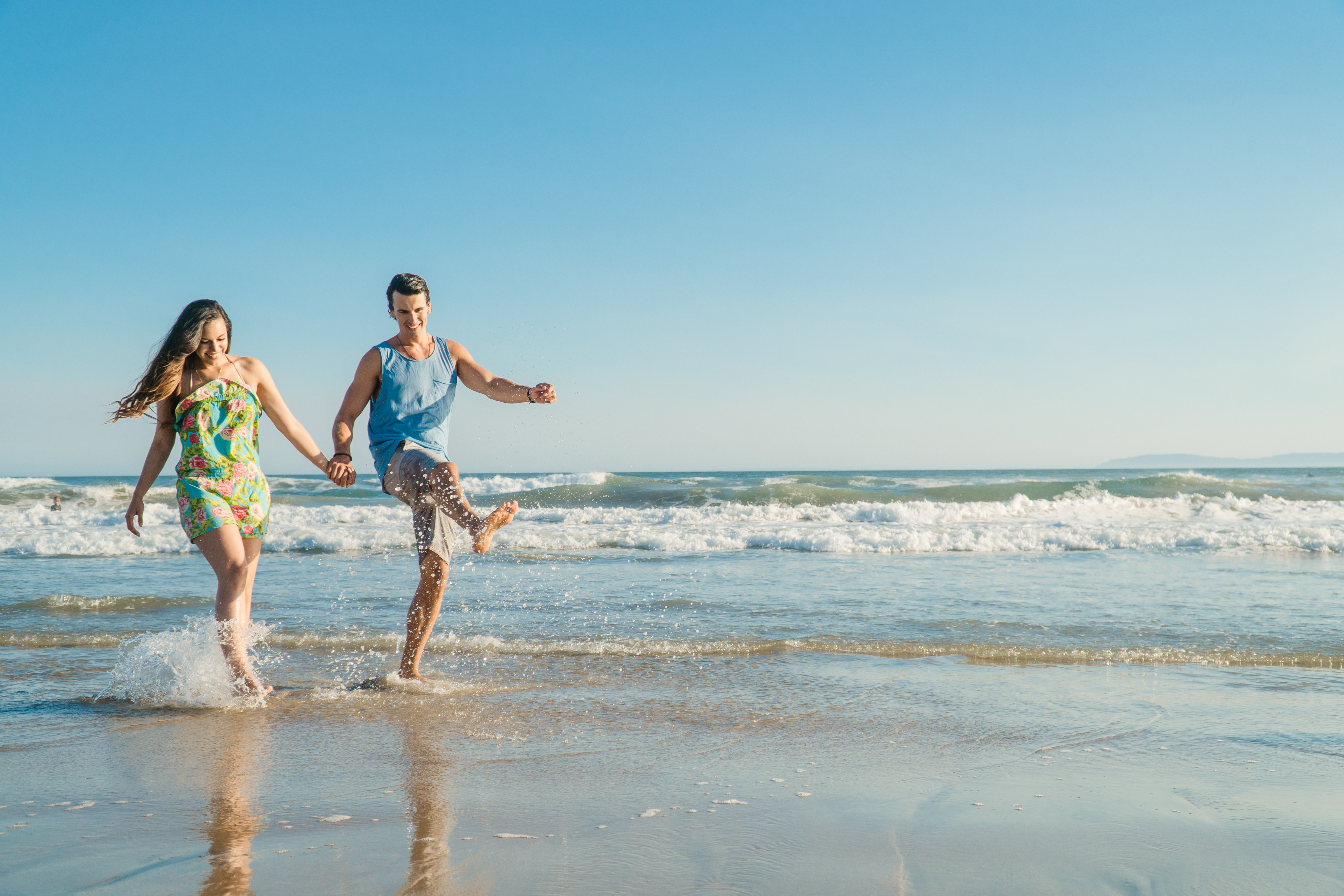 A couple joyfully holds hands and splashes in shallow ocean waves on a sunny Huntington Beach, California day, with a clear blue sky and distant coastline in the background.