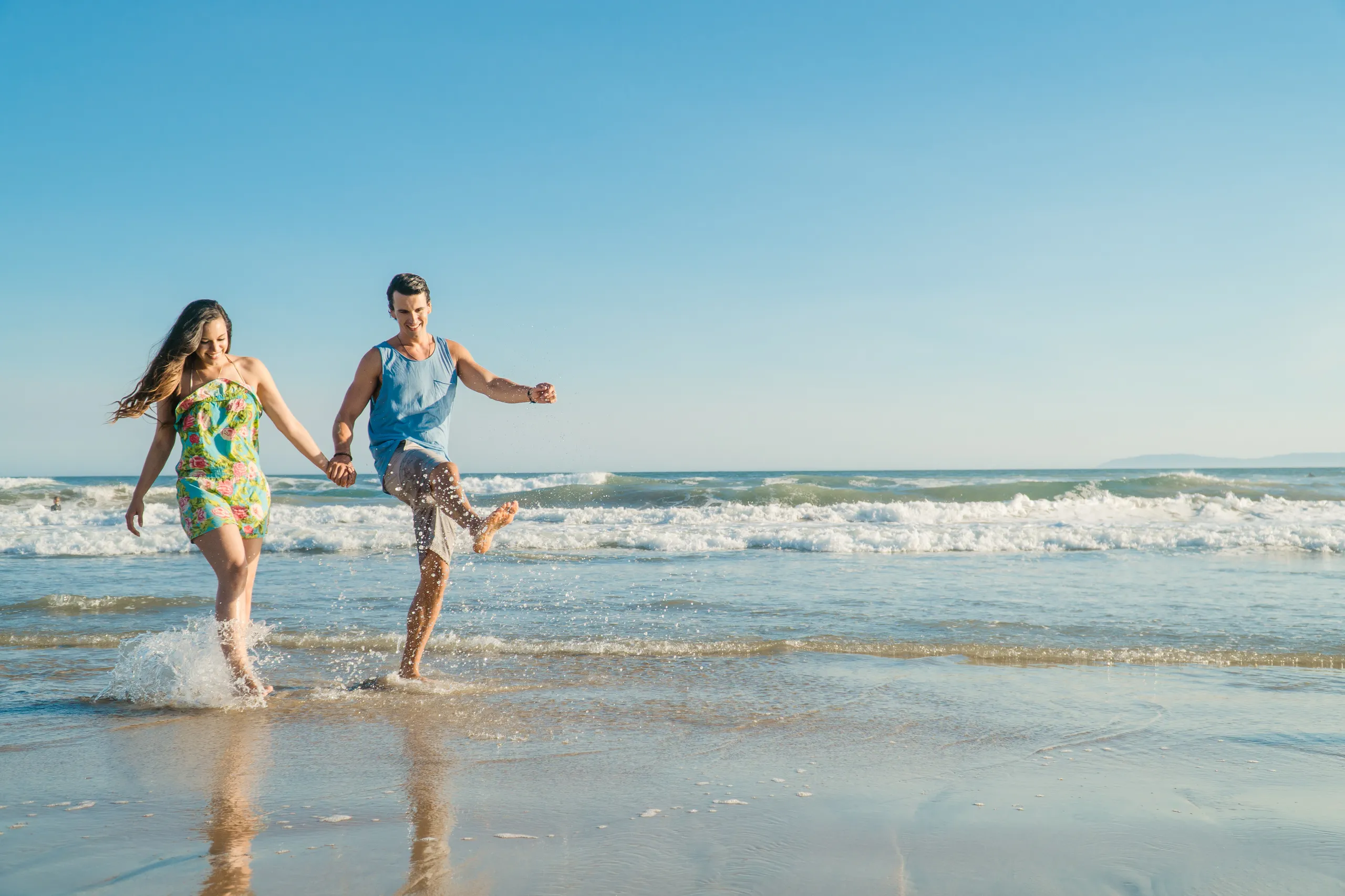 A couple joyfully holds hands and splashes in shallow ocean waves on a sunny Huntington Beach, California day, with a clear blue sky and distant coastline in the background.