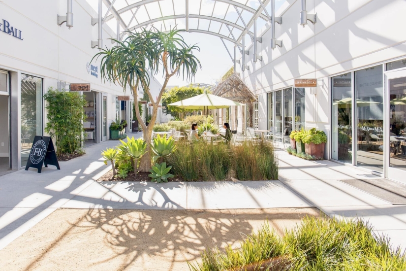 A bright, modern outdoor shopping area in Costa Mesa with plants, palm-like trees, and tables with umbrellas. Glass storefronts line the walkway as sunlight filters through a curved glass roof, casting soft shadows on the ground.