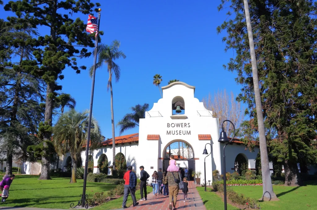 Visitors walk toward the white, Spanish-style entrance of the Bowers Museum, just a short drive from Costa Mesa, surrounded by tall palm and pine trees, green lawns, and a clear blue sky. An American flag flies on a flagpole to the left.
