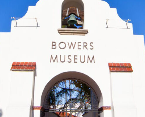 Visitors walk toward the white, Spanish-style entrance of the Bowers Museum, just a short drive from Costa Mesa, surrounded by tall palm and pine trees, green lawns, and a clear blue sky. An American flag flies on a flagpole to the left.