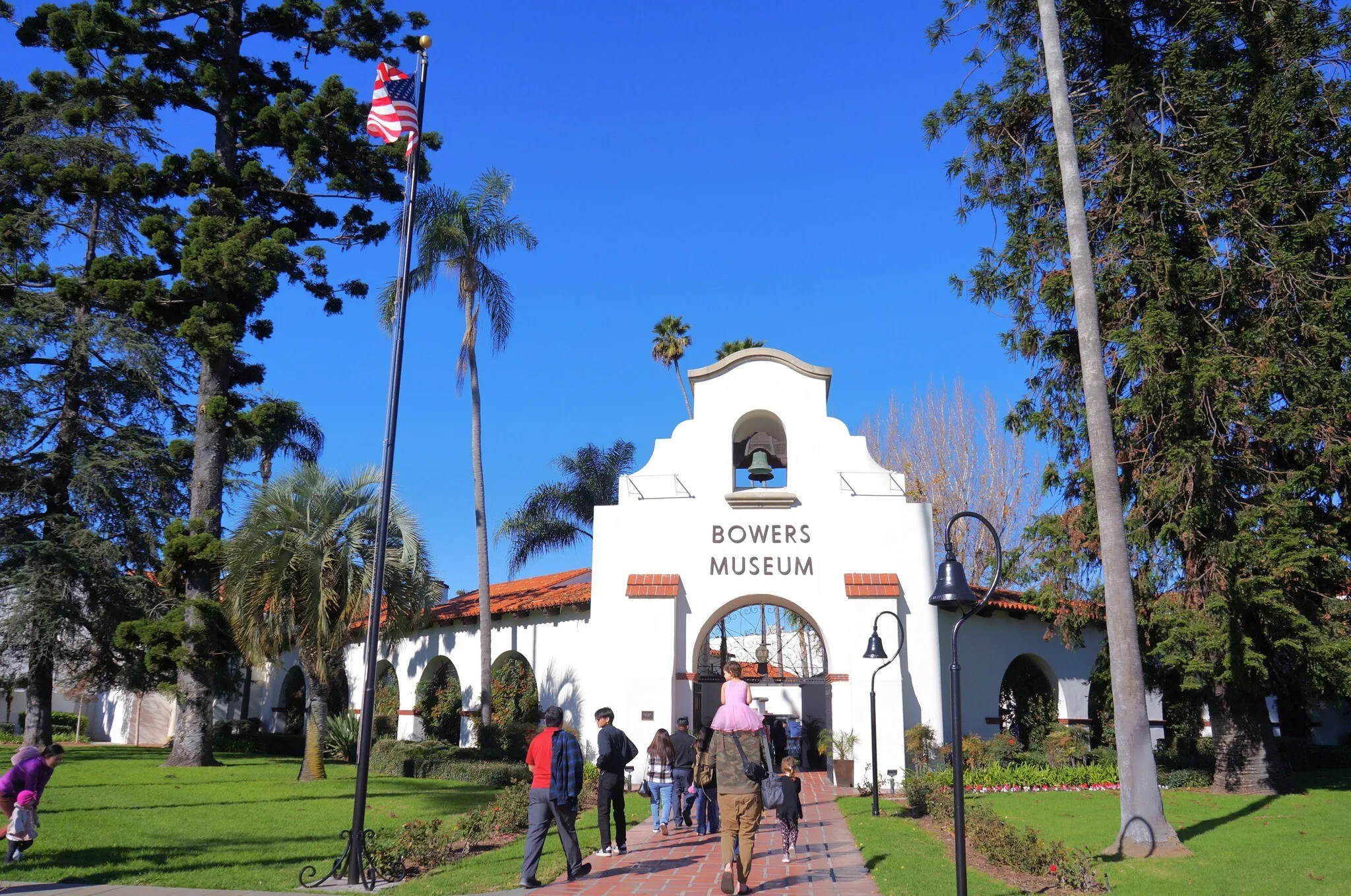 Visitors walk toward the white, Spanish-style entrance of the Bowers Museum, just a short drive from Costa Mesa, surrounded by tall palm and pine trees, green lawns, and a clear blue sky. An American flag flies on a flagpole to the left.