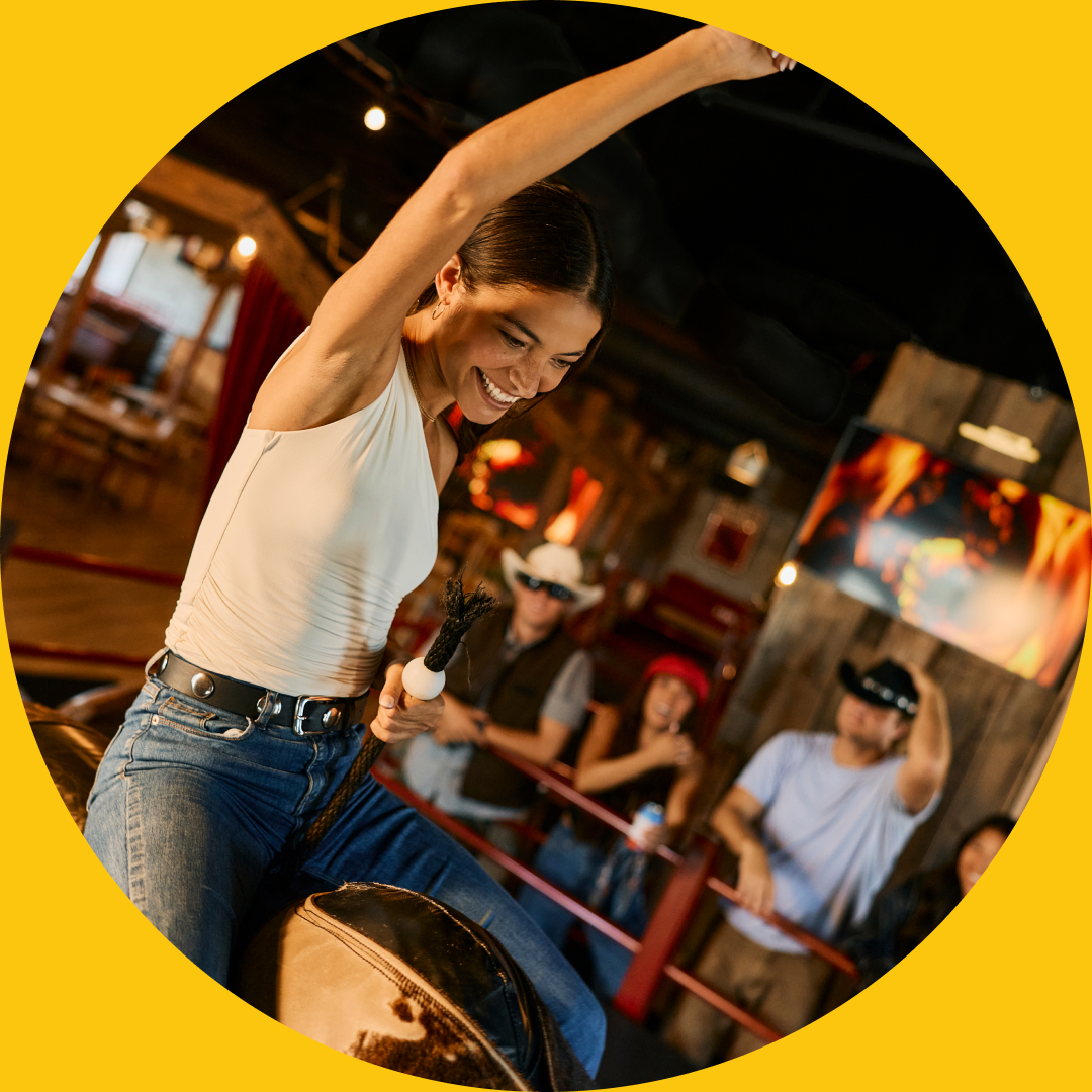 A smiling woman wearing a white top and blue jeans rides a mechanical bull indoors in Costa Mesa, raising one arm as a group of people watch and cheer. The image is framed by a yellow circular border.