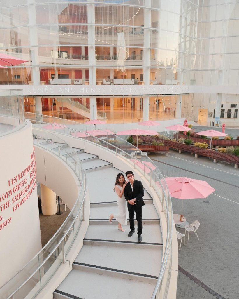 A couple dressed in formal attire stands on a curved white staircase outside a modern glass building with pink umbrella-covered tables on the patio below.